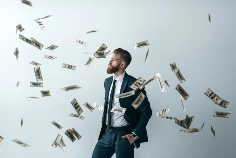 Bearded man in a dark business suit standing confidently with his hand in his pocket while US dollar bills rain down around him against a gray background. This illustrates the potential for wealth accumulation and passive income through high-yield savings accounts, which are currently offering interest rates up to 4.15% APY as of July 2026, allowing savers to maximize returns despite fluctuating market conditions. Source: The College Investor