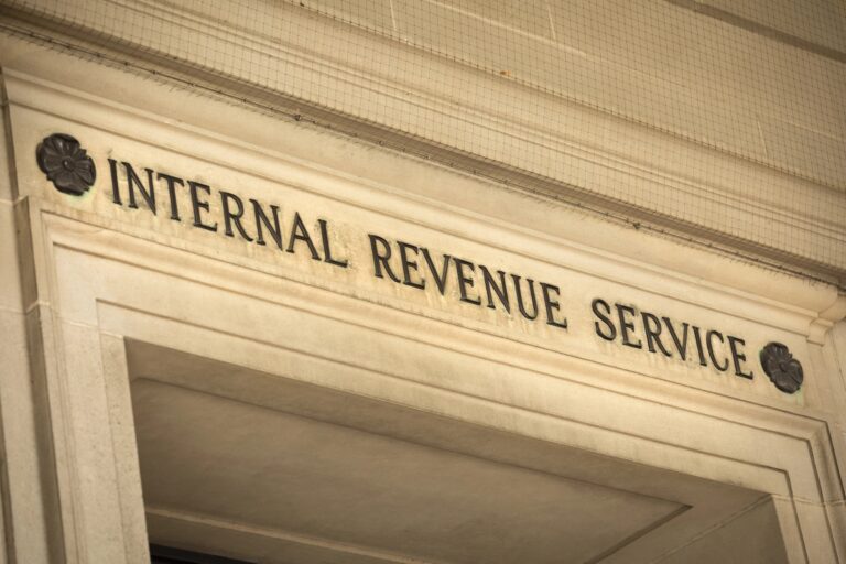A close-up, slightly upward-angled shot of the Internal Revenue Service (IRS) federal building's entrance in Washington D.C., USA, emphasizing its prominent stone facade and the bold, black carved letters spelling "INTERNAL REVENUE SERVICE" above the doorway. The classic, ornate architectural details, including decorative dark rosettes flanking the text and intricately carved stone trim, are clearly visible. The image conveys a sense of official authority and directly relates to the article discussing the IRS's annual inflation adjustments for retirement accounts, specifically the raised 2026 contribution limits for 401(k) and IRA plans, as detailed in Notice 2025-67.