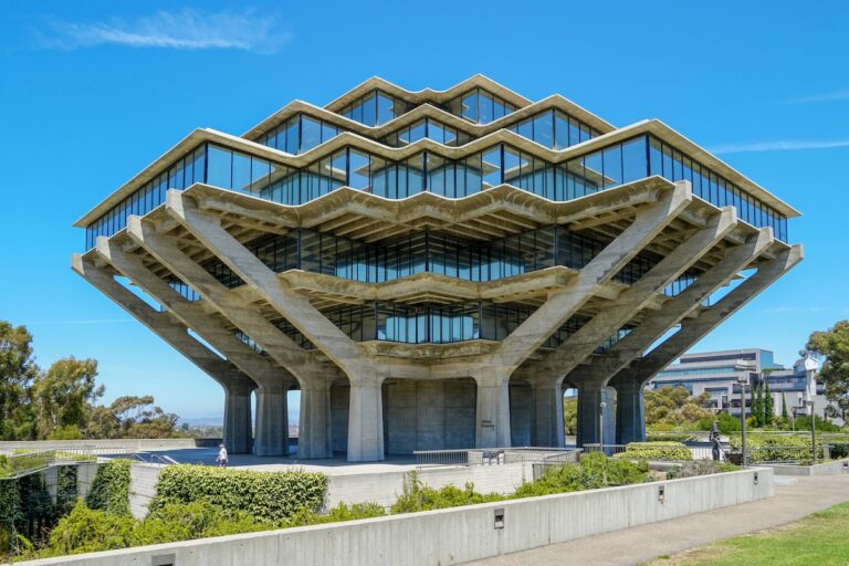 The iconic Geisel Library at the University of California San Diego stands majestically against a bright blue sky, its unique brutalist architecture resembling hands holding up a stack of books. This striking eight-story concrete and glass structure, with its geometric patterns and expansive windows reflecting the surroundings, is framed by lush green landscaping and pathways, hinting at the vibrant campus environment. The image visually anchors the article discussing concerns about UCSD freshmen's declining math skills and the university's response with remedial courses, raising questions about admissions practices, test-optional policies, and grade inflation amidst record demand for college admissions.